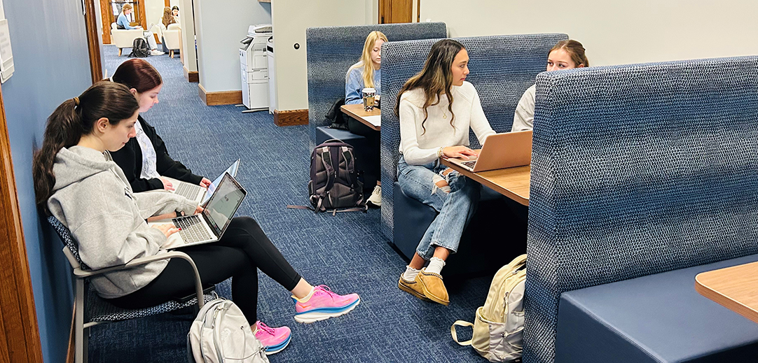 multiple female students in booths and in chairs talking and looking at their laptops