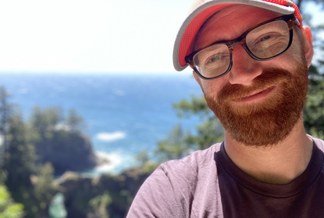 Daniel Meyers with beard, glasses, cap, pink shirt with the ocean in background