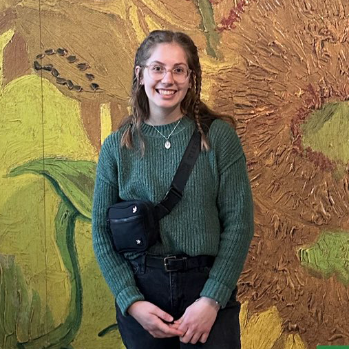 girl with brown hair in braids, round glasses, green sweater, in front of van gogh's sunflowers painting