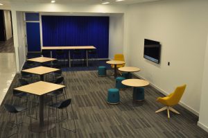 A collaborative workspace in the new Sciences Complex, filled with a variety of small tables--some high, some low-- along with chairs, padded stools, and a screen for presentations on the wall