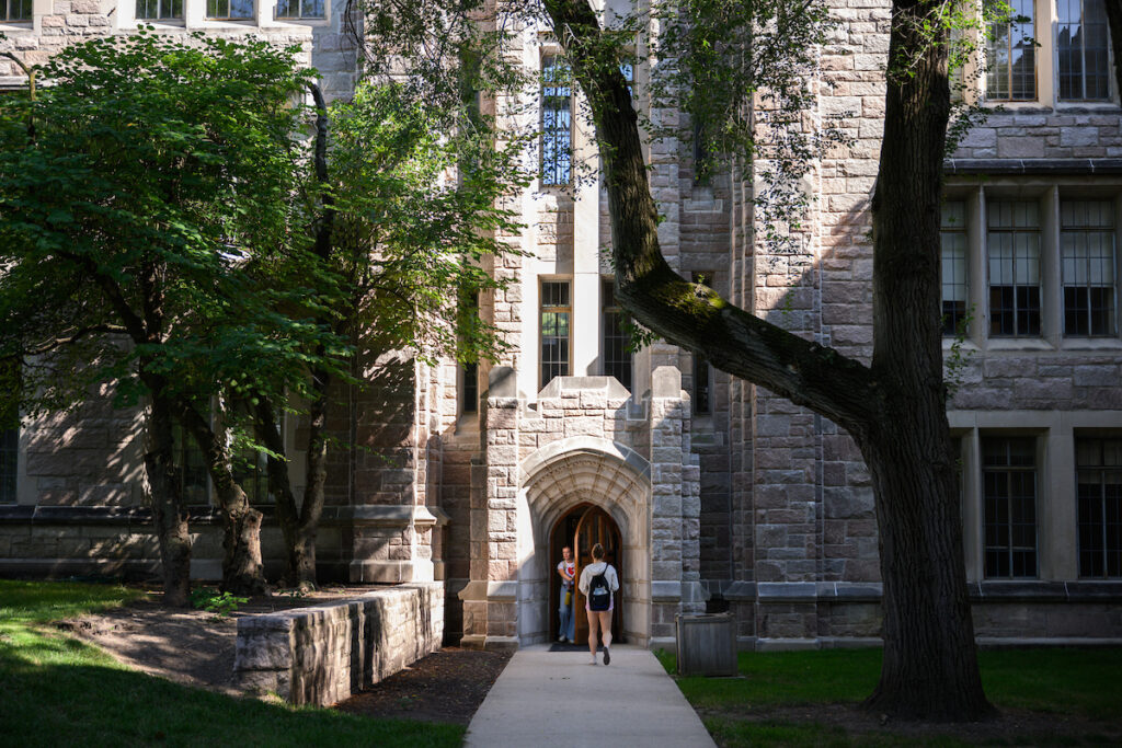 limestone building shaded by green trees, student with backpack entering through wooden door