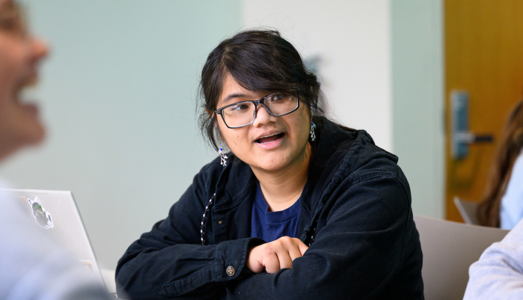 female in blue sweatshirt, with dark hair and dark-rimmed glasses