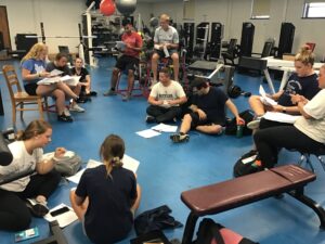 A classroom of athletic trainers looks at sheets of paper in a weight room. 