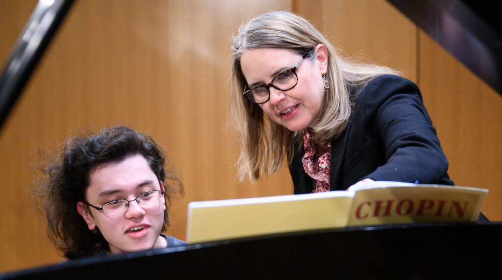 female faculty in blue blazer and glasses at piano with male student with dark hair and glasses