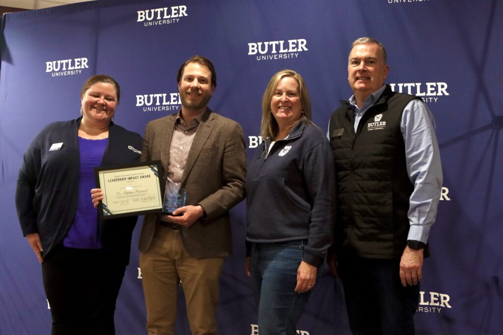 Dr. Stephen Barnard poses with his certificate and trophy alongside Director of New Student & Family Programs Meg Haggerty (left) and nominating family Jamie & David Fryrear (right). Butler University backdrop with four people posing in front. Dr. Barnard holding his certificate and trophy.