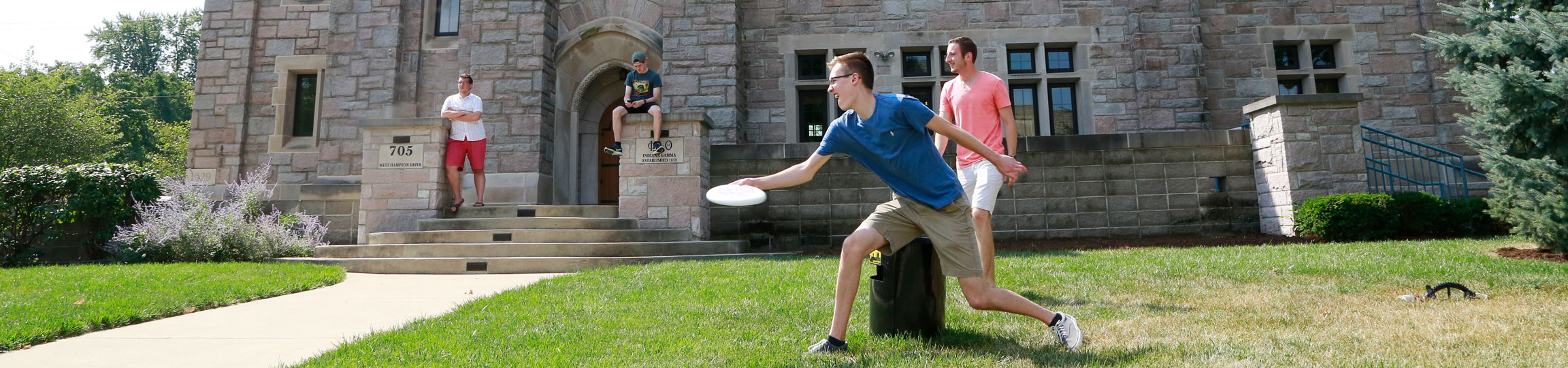 Students play frisbee in front of a campus building