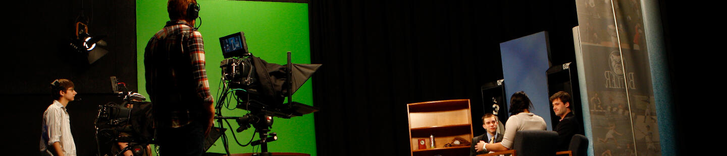 green screen on wall, tv cameras and cameramen, 2 males and 1 female talking on stage
