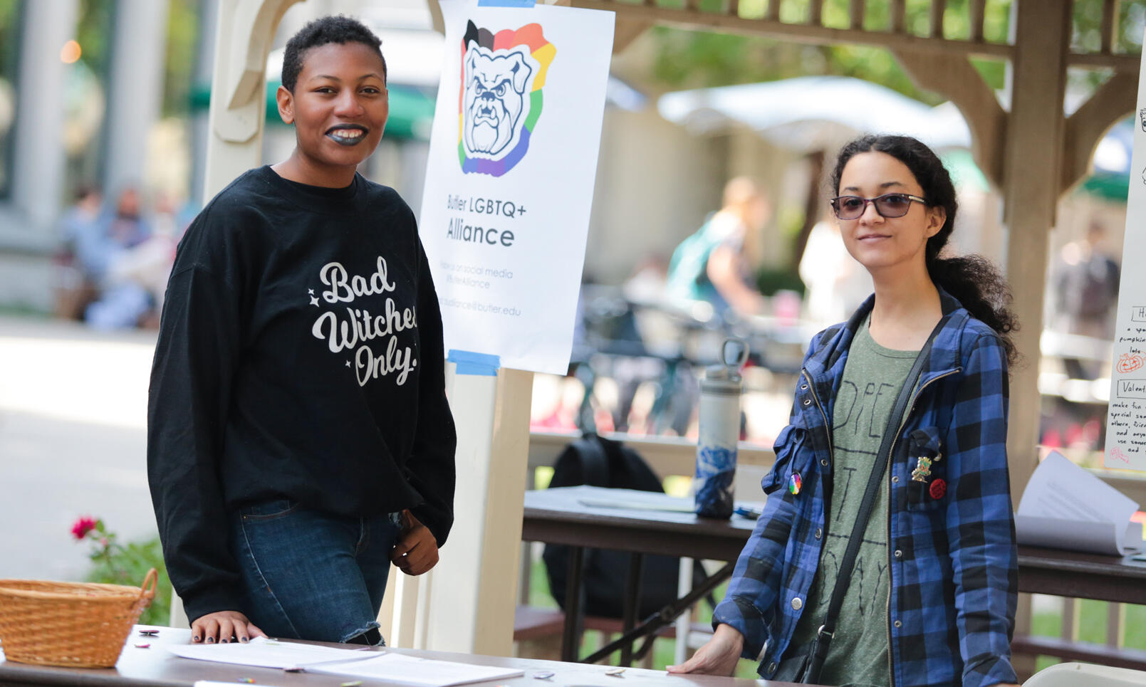 two LGBTQ+ Alliance members with that signage, one in a black sweatshirt reading Bad Witches Only, the other with sunglasses and an blue and black plaid shirt