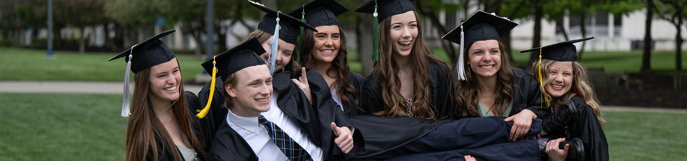 6 female students in graduation wear holding up one male in graduation wear